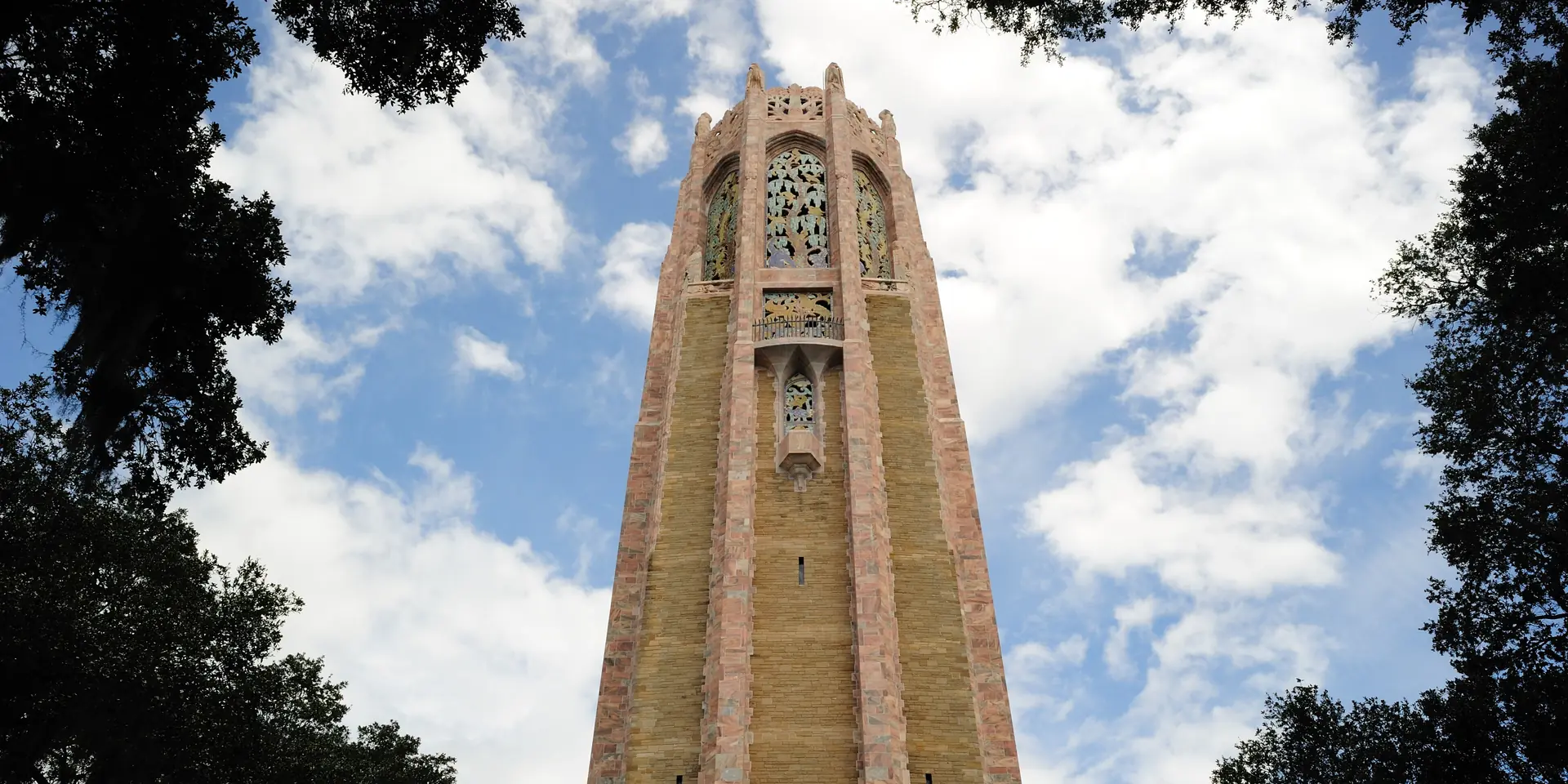 Bok Tower in Lake Wales seen from below through the trees, pink and gray stone against a blue sky with clouds